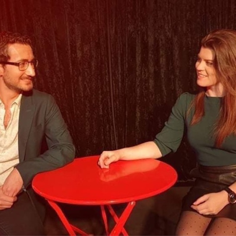 Benjamin and Veronika sit at a red table, as if on a date. They are looking at each other and smiling