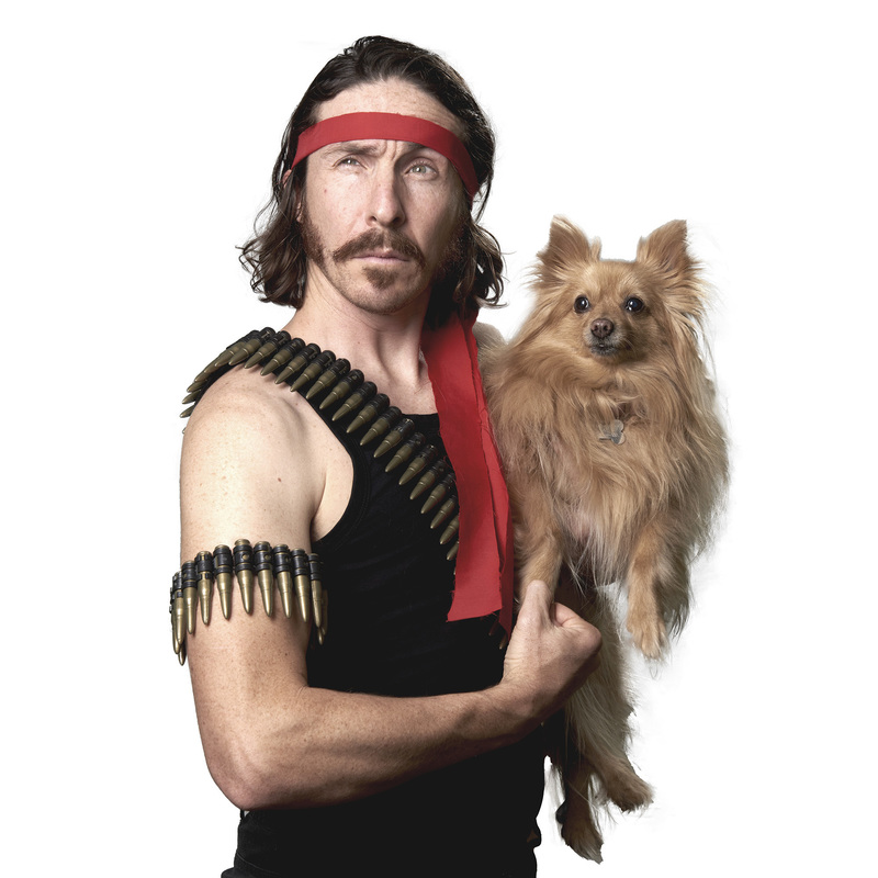 Man looking serious yet silly with a red headband on his head, wearing a black singlet with a bullet belt slung over his shoulder, felxing his right arm, which also has a small bullet belt wrapped around his bi-cep. In his left arm he holds a cute, light brown fluffy dog.