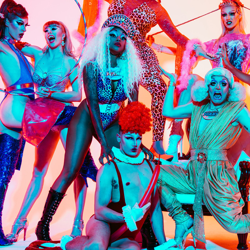 YUMMY: ICONIC - Six drag burlesque performers dressed in revealing and colourful clothing pose in front of a yellow and pink wall.