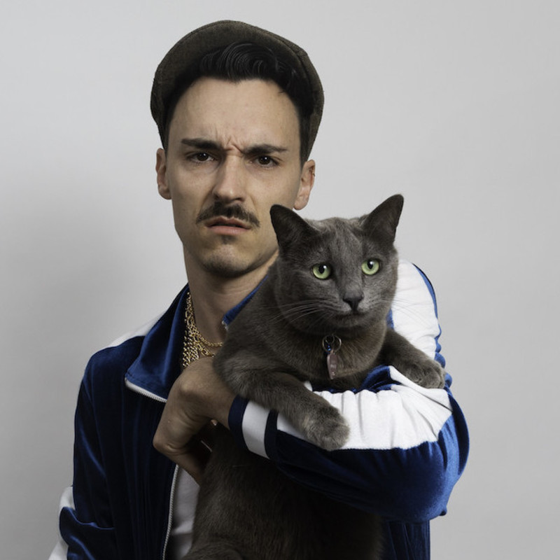 Comedian Andrew Silverwood is wearing a blue tracksuit top and a white shirt and is holding a grey cat. They both look concerned.