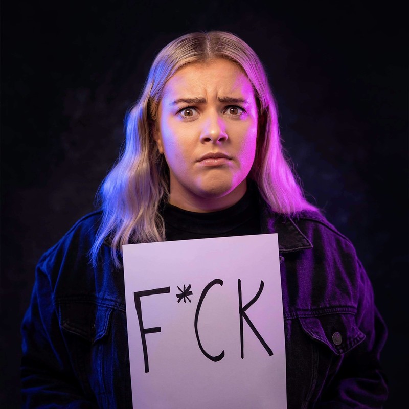 Taylor Nobes against a dark background holding a sign saying "F*CK"