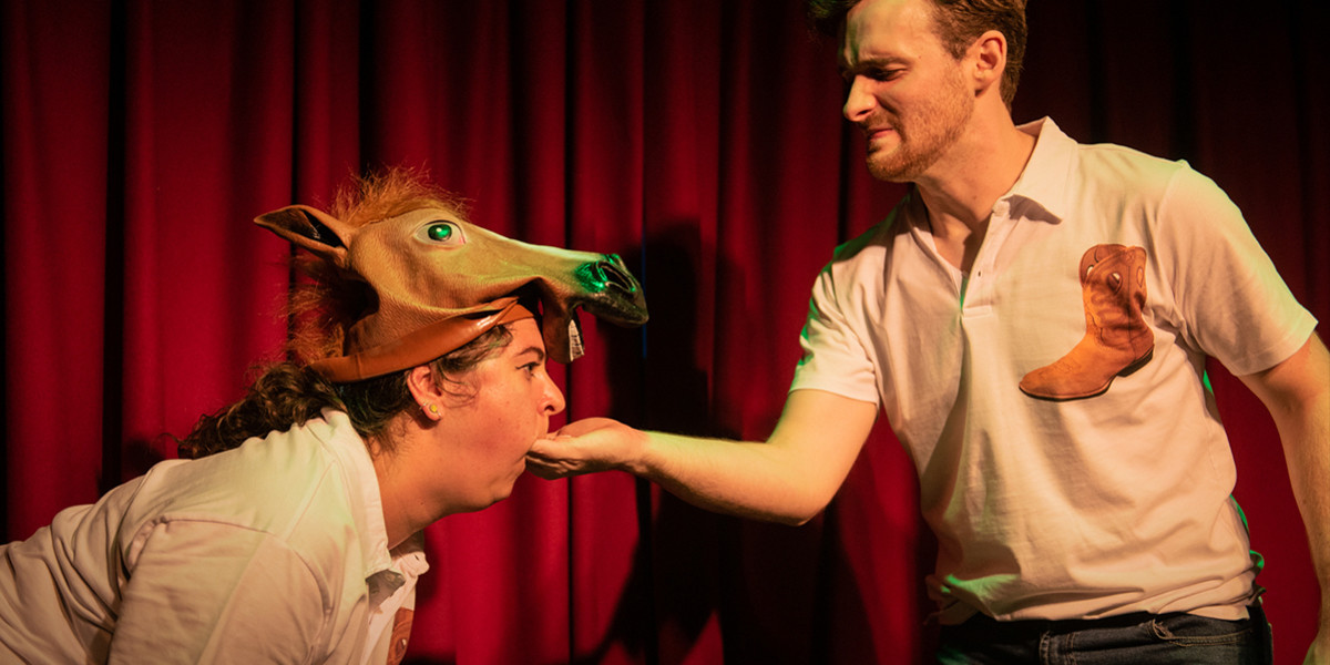 Doug Rintoul, dressed in a white boot-themed polo shirt, holds his hand out towards Katie Currie, who has put it in her mouth in its entirity. She's wearing a rubber horse mask on top of her head. Doug looks extremely displeased with the situation.