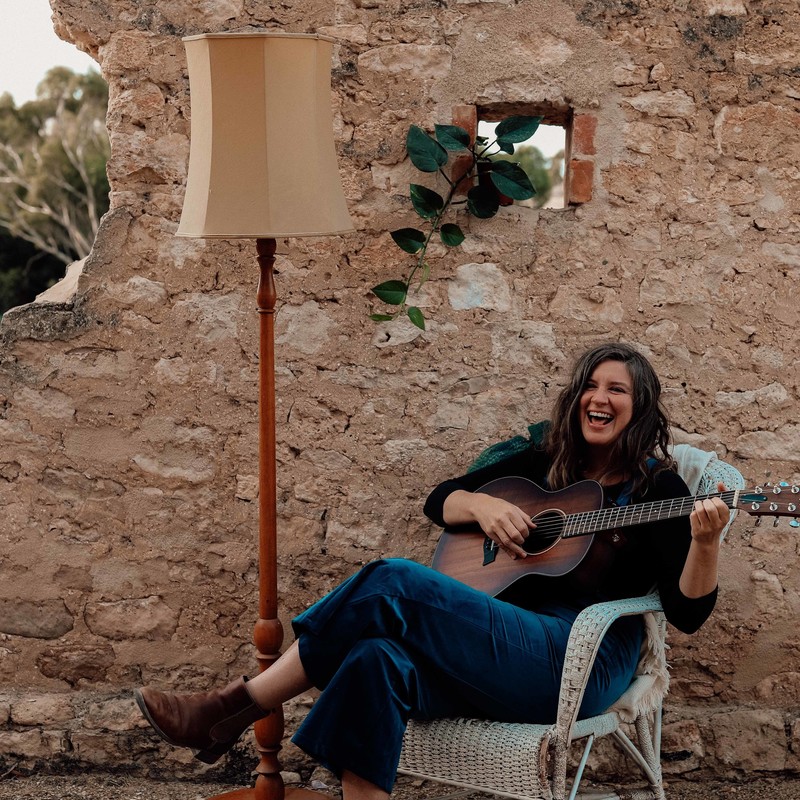Woman sits on chair in front of limestone wall. She holds a dark brown guitar. a tall lamp stands next to her and a pot plant behind.