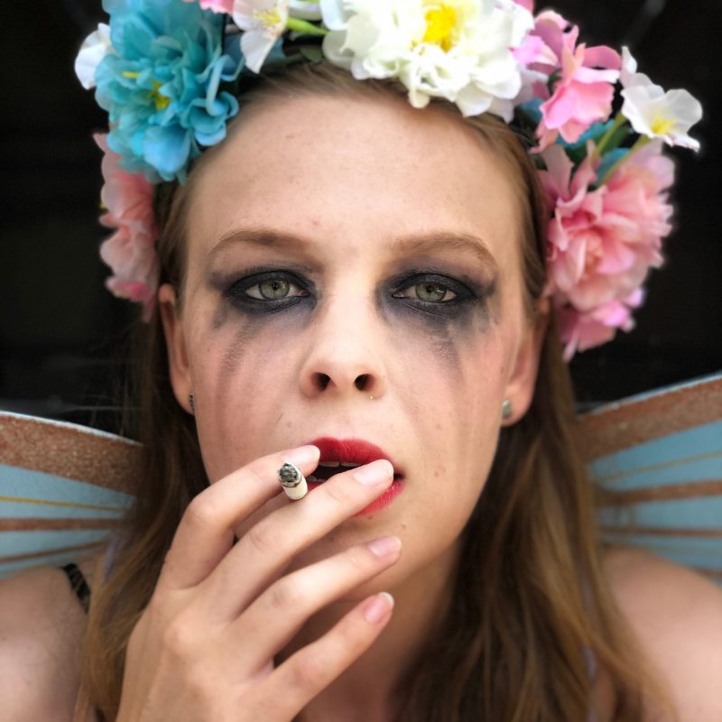 A woman in a flower crown and fairy wings, with smudged dark makeup, looks at the camera while smoking a cigarette