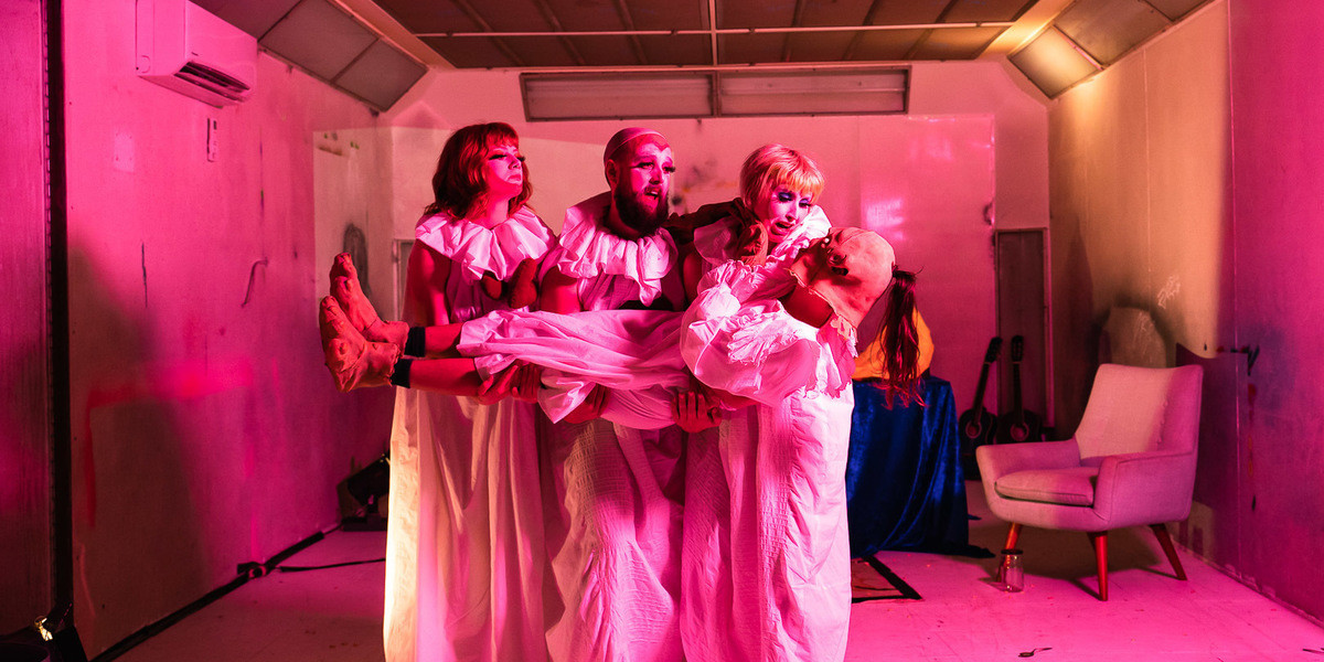 Three figures in white nightdresses and clown makeup stand in a line and hold someone in a ghost costume length ways in their arms.