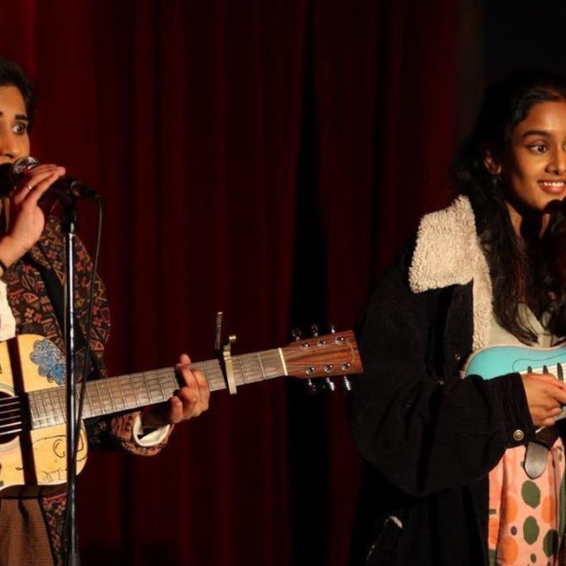 Leela and Shabana are performing a set at Rhino Room. The Rhino room sign is on the right hand side of the image on the wall. Behind them is a dark red, velvet curtain. They both have microphones on stands in front of them and one in front of Leela's guitar. Leela's guitar is a wooden, acoustic guitar with drawings on it of a rainbow and elephants and monsters. Leela is holding the microphone and wears a brown and red patterned jacket. Shabana is smiling and wears a black oversized jacket with sheep wool on the inside.