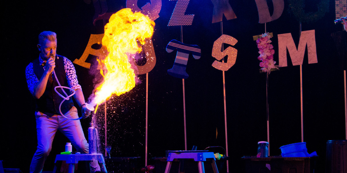 A man with silver hair and a grey beard is blowing cornflour through a pipe to create a large fireball. Colourful letters on long silver poles are behind the man.