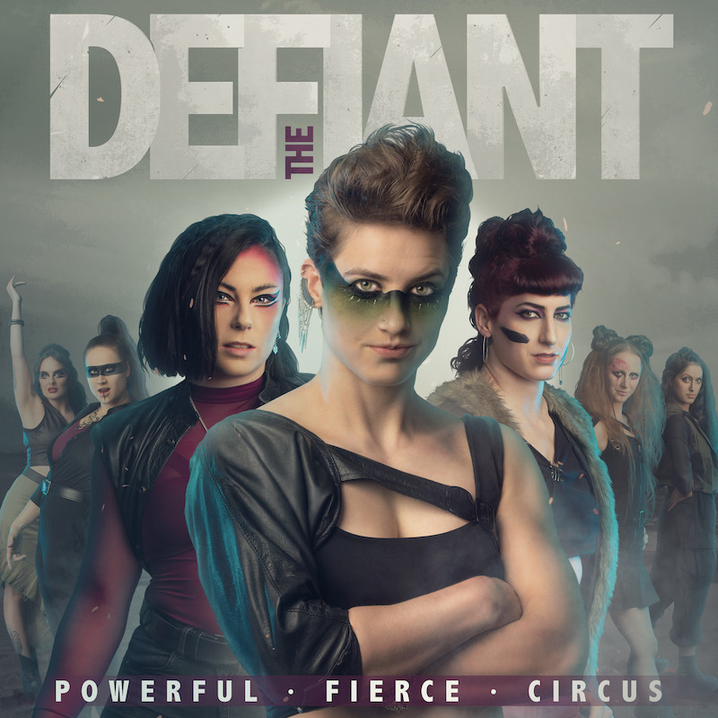 The Defiant - The Defiant. Powerful. Fierce. Circus. Seven women and non-binary artists stand facing the camera in distressed clothes and warpaint and make up with a post-apocalyptican background.