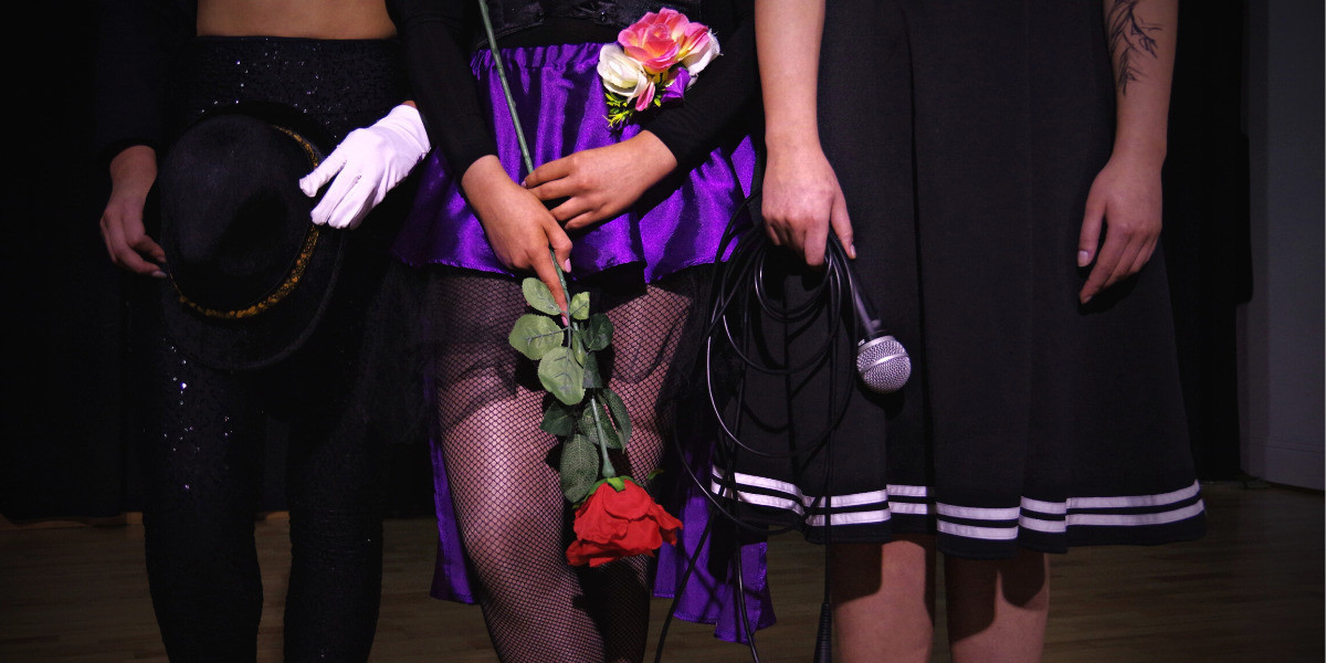 A picture of the bottom half of 3 girls. One wearing black pants and holding a black fedora, one wearing a purple skirt holding a rose and one in a black dress holding a microphone.