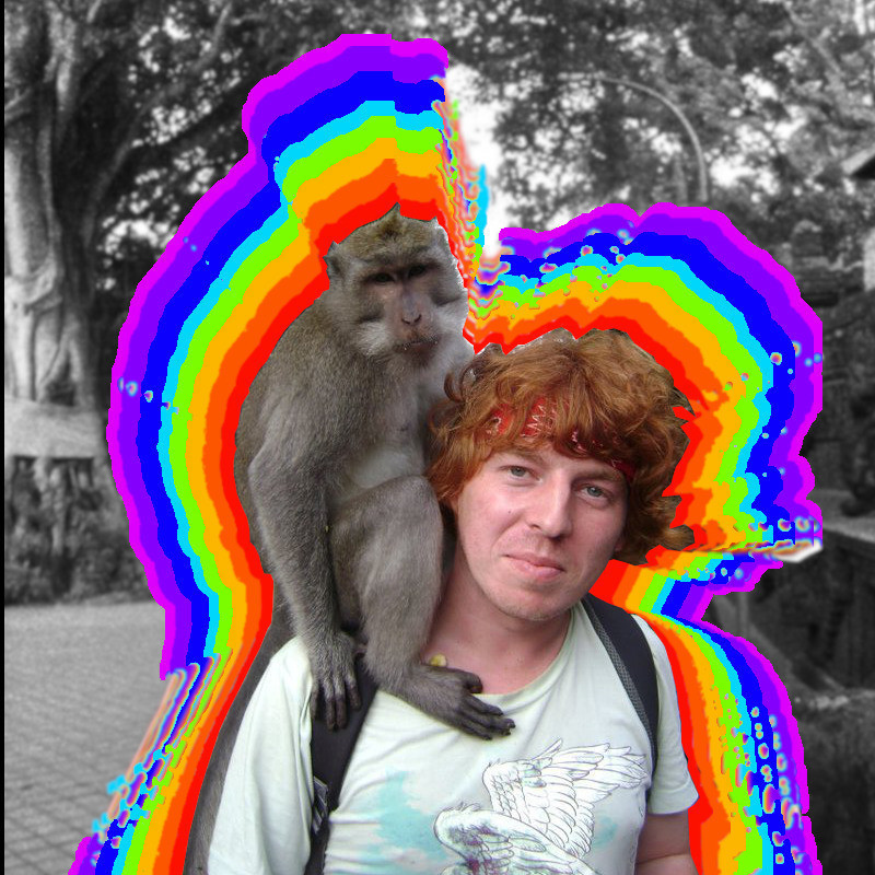 A monkey sits on the shoulder of a man with short red hair, he wears a red bandanna and a light blue t-shirt. A rainbow radiates from them creating a border. The background is black and white trees and a footpath.