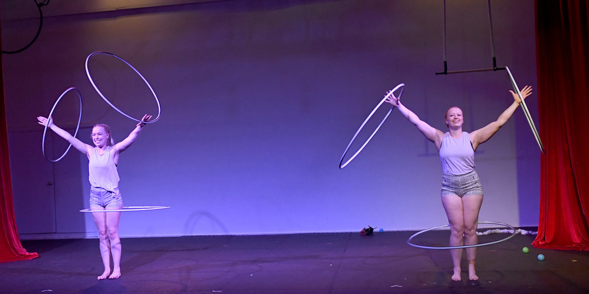 2 females, Zoe and Alex hula hoop 1 hoop around their knees and 2 in each hand. They are on a stage with red curtains and the lighting is bluey/purple.