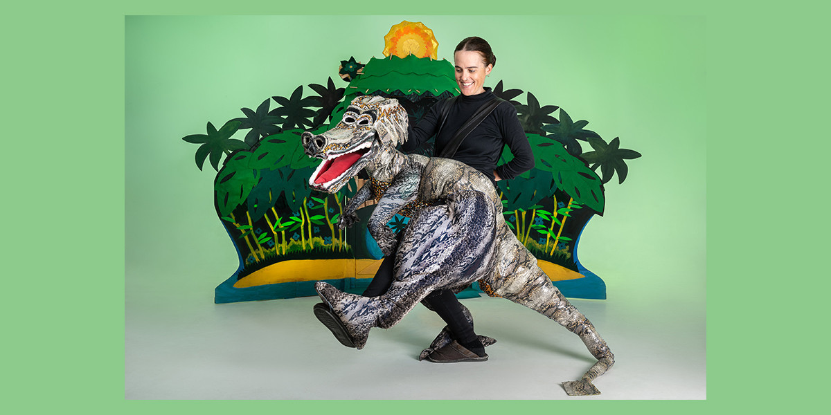 The puppeteer stands inside a dragon puppet with its leg kicking in a dance move. Behind them is a set of a tropical island.