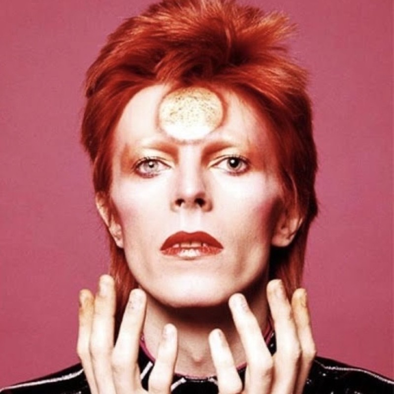 Five Years (1969-1974) - Ashes To Ashes Bowie Tribute - A head shot of David Bowie. He has fair skin, bright red lipstick, gold eyeshadow and spiky red hair. He has a gold circle painted on his forehead. The background is pink.