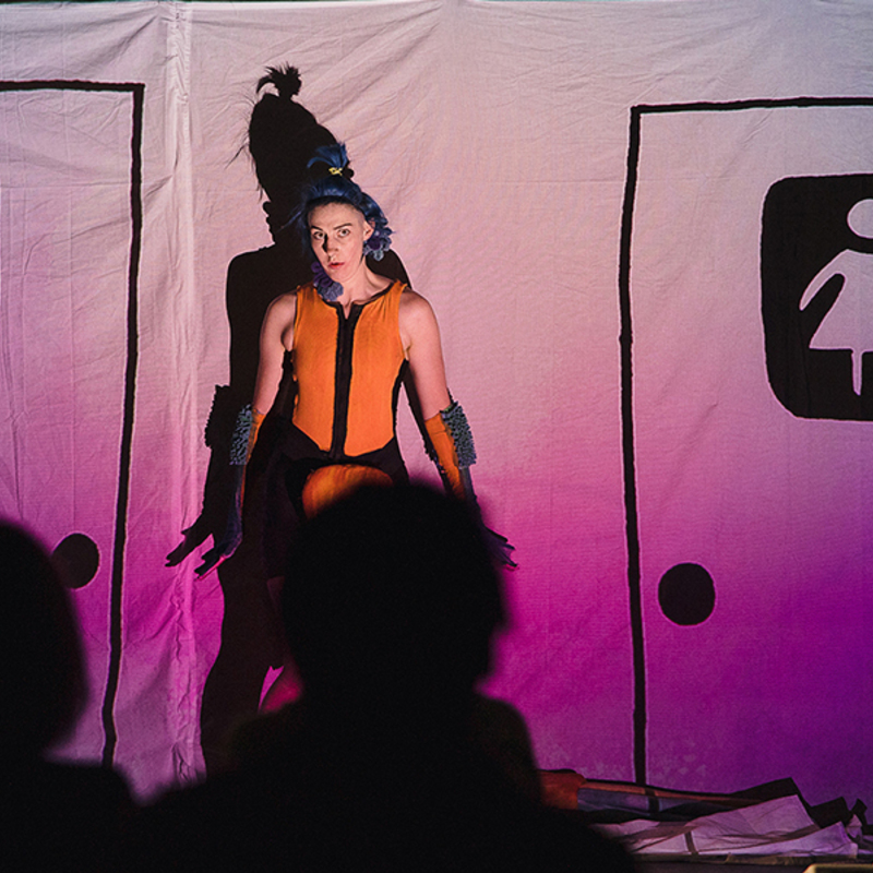 A non-binary performer in an orange bathing suit stands in front of a screen that has a hand-drawn projection of gendered bathrooms stalls.