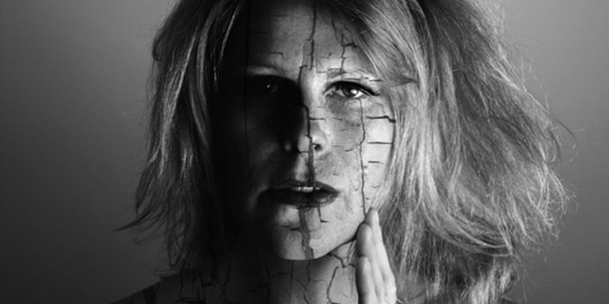 A black and white image of a woman's face with her light coloured hair flowing behind her. Her left hand is touching the side of her face and she looks intensely at the camera. Her face has artificial cracks on it.