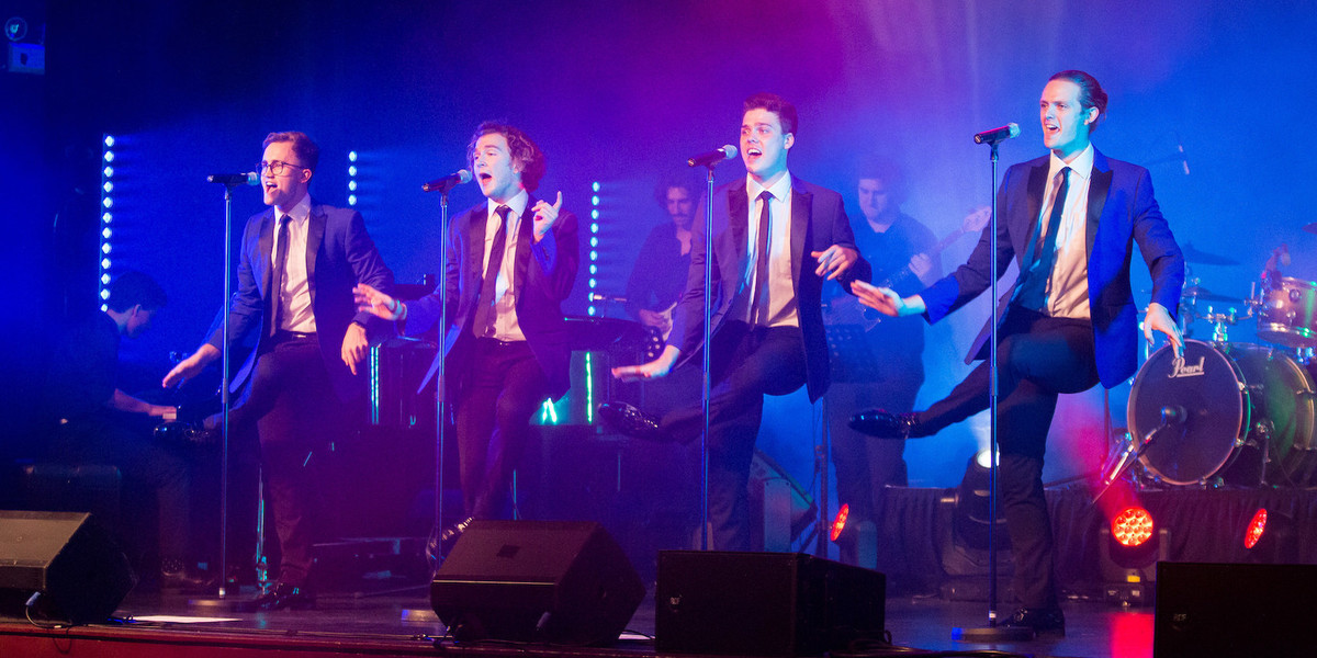 Four men are on stage, facing the audience in a line behind four microphones on straight stands. They are dancing and singing, with their left knee being hit by their right hand in a 60s-style disco move. In the background, a live band, including a pianist, a guitarist, a bassist and a drummer. The lighting create blue ambience, with red floor lighting creating a purple haze around their heads.