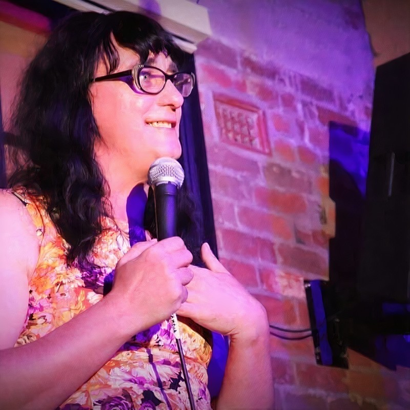 A photo of a person holding a microphone. They have long black hair and they are wearing black framed glasses and floral top.