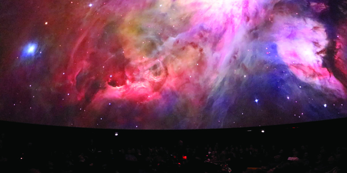 Musicians sits in the round surrounded by a full audience gazing up into a planetarium dome. The dome ceiling features the glow of stars through dark cloudy spirals of purple, blue, amber and pink.