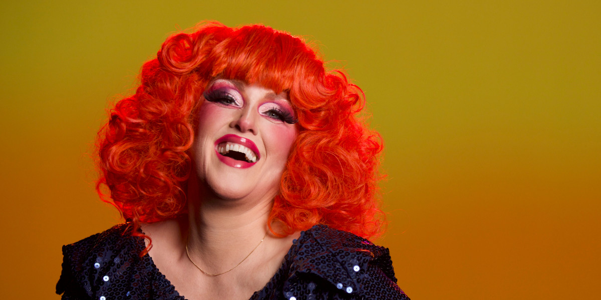 Woman in drag make-up with bright orange hair throws back her head to laugh, wearing a blue sequinned dress, standing against a golden background. Photograph by Evan Munro-Smith.