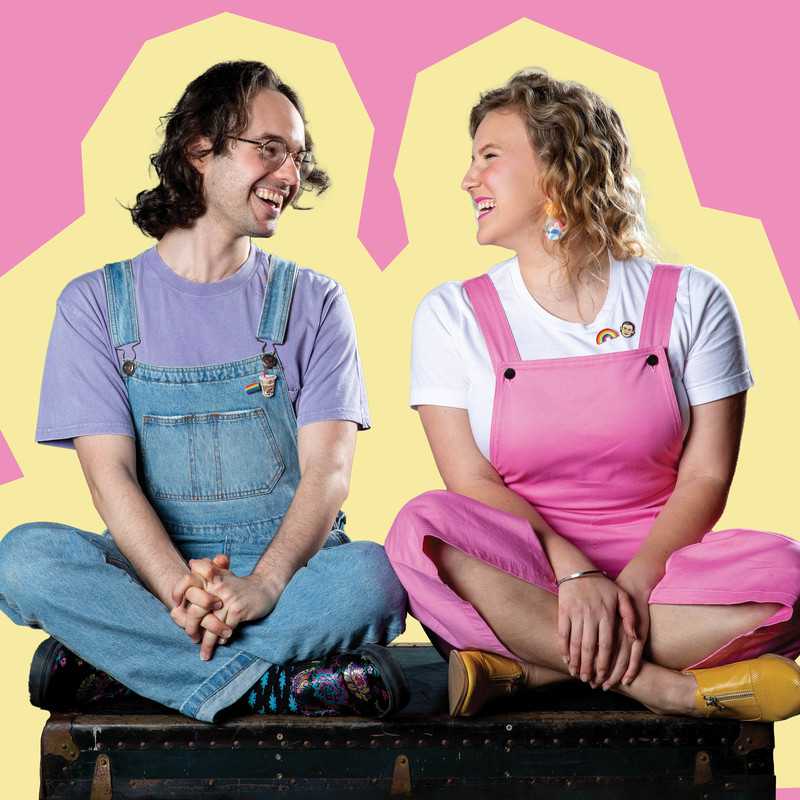 A photograph of two people smiling at each other. The person on the left has medium length brown hair and is wearing clear glasses. They are also wearing a light purple t-shirt with denim overalls on top. Their shoes and socks are patterned and colourful. The person on the right has medium length blonde curly hair they are wearing a white t-shirt with pink overalls and small yellow boots.
