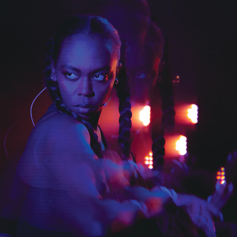 A black woman with long braided hair stands side on. Her head is turned, looking seriously as something just past the camera. A blue filter is across the image