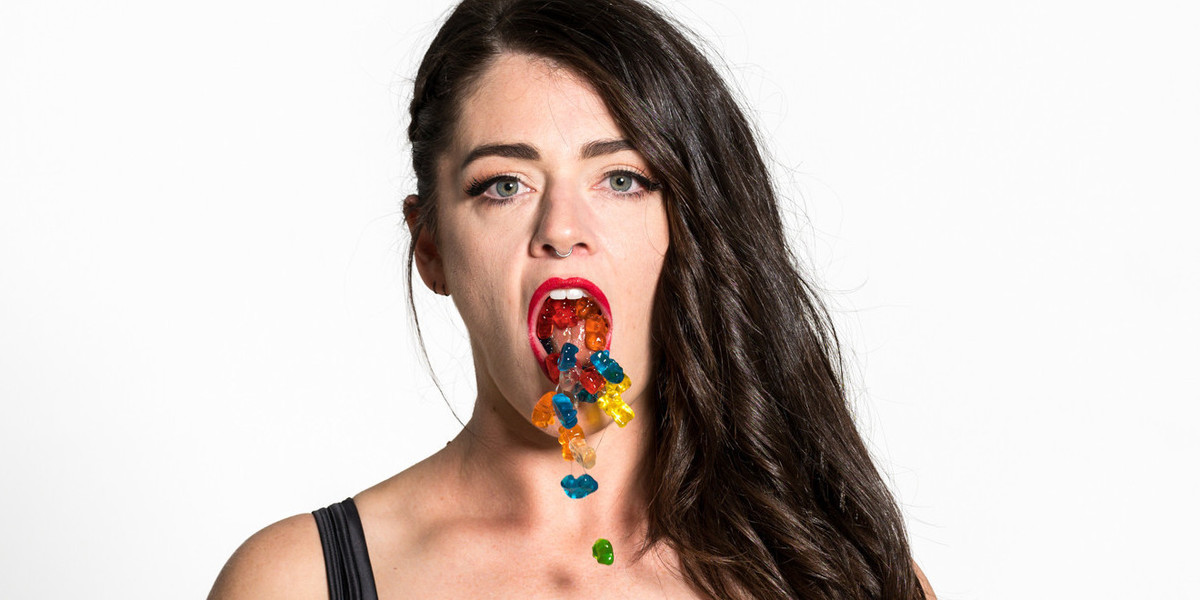 A girl with long brown hair spits gummy bears from her mouth on a white background.