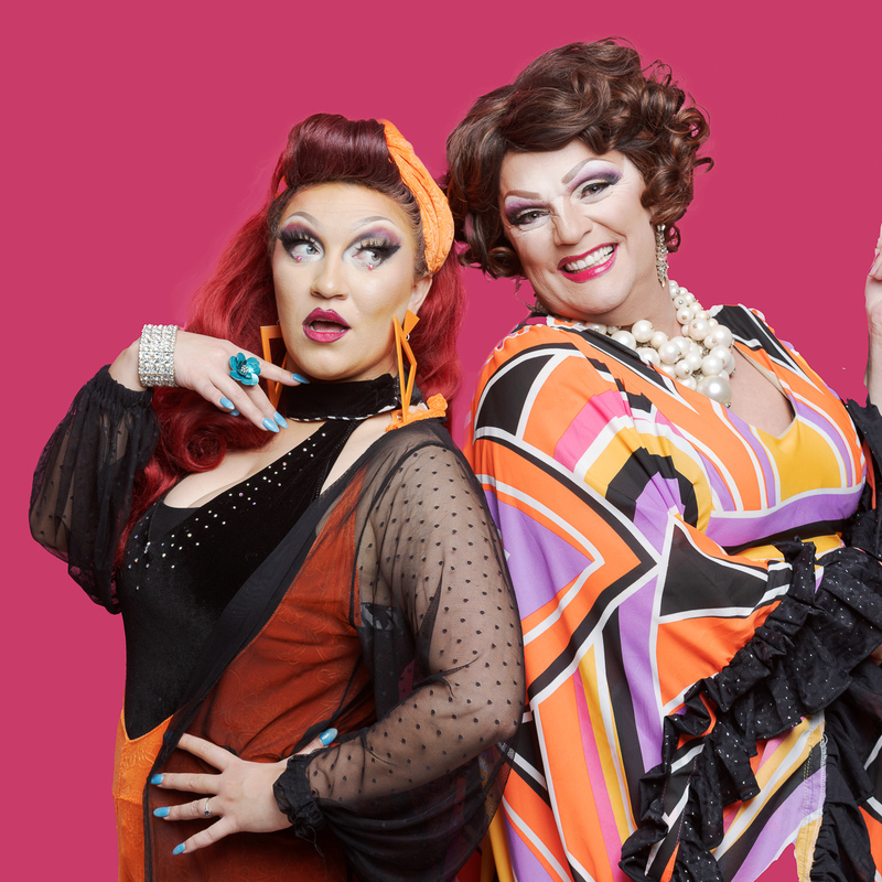 Two drag queens stand back to back on a pink background. They are both brightly coloured in orange and black clothing.