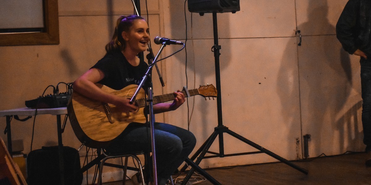 Poppy sits on a chair in a hall, to the left of centre of the image. She is wearing blue jeans and a black tshirt. She holds a guitar on her lap and is singing into a microphone. She is smiling and looks happy. A speaker on a stand sits to the right of the image.