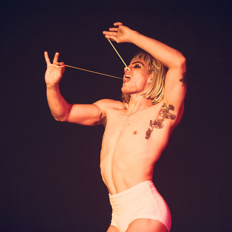 A performer in a blonde wig and white underwear with a long balloon threaded through their nose and out their mouth holding it stretched with their hands.
