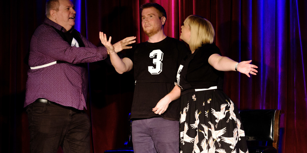Three improvisers performing on stage wearing netball bibs. Each bib has a number on it. One improviser looks out and gestures to the crowd.