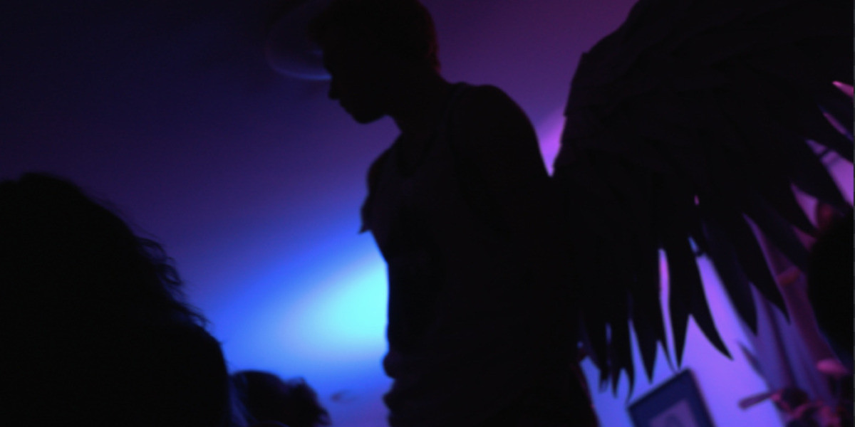 The silhouette of a young man with large wings on his back, standing above a crowd of people.