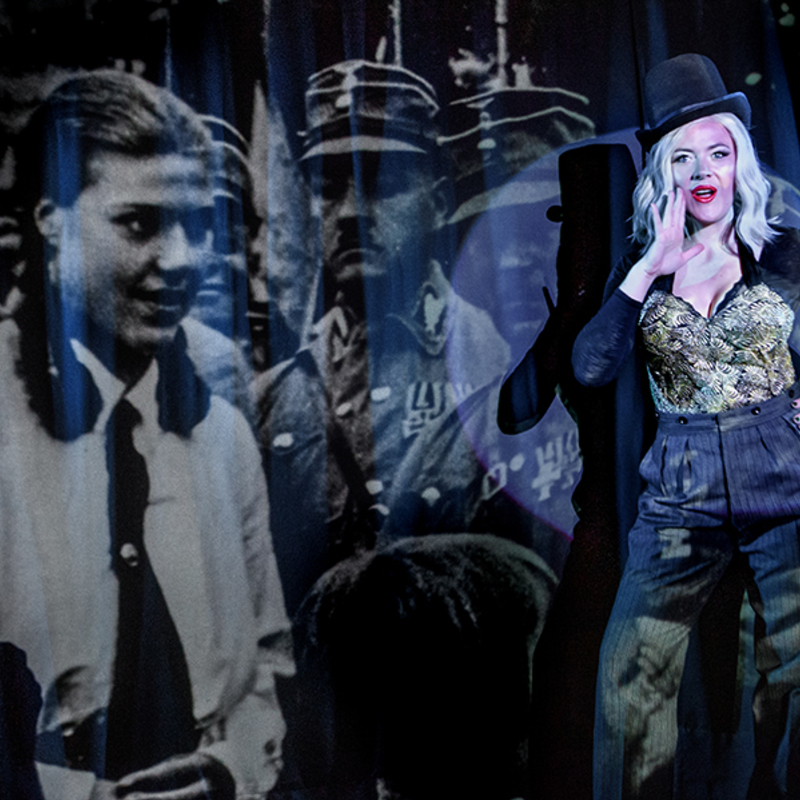 A cabaret performer is gesturing a secret with her hand on her hip, whilst standing in front of a projection of a german female with Nazi troopers. The cabaret performer is wearing a top hat, golden bodice and trousers, and she has white hair and red lips.