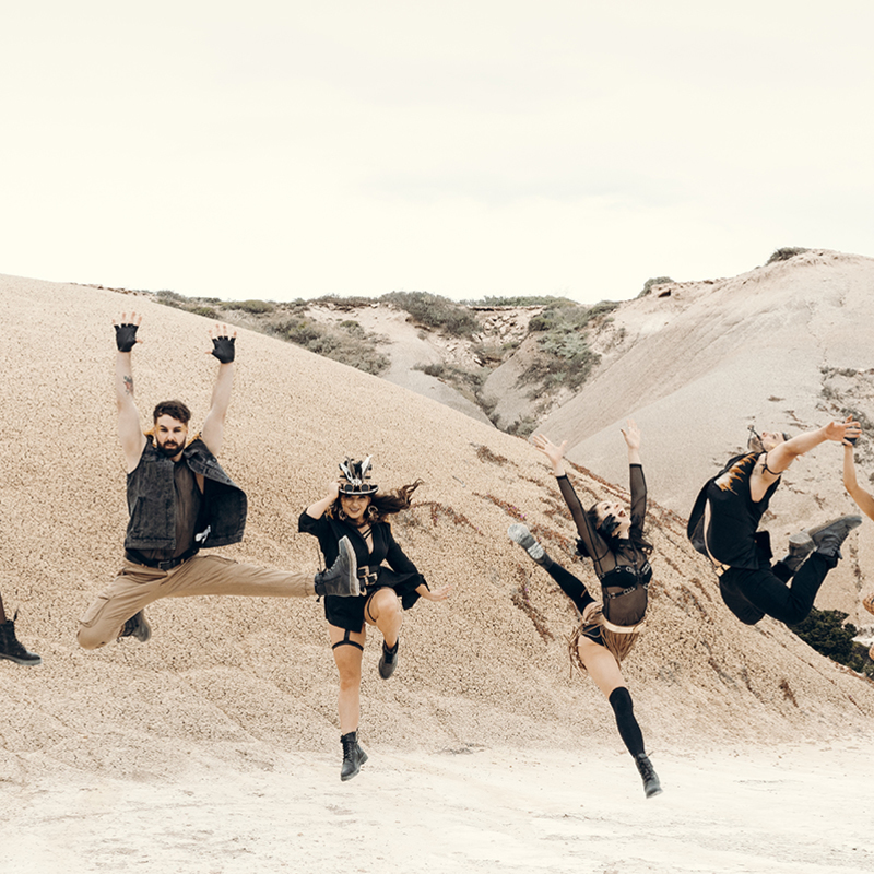 A desert scene with 6 people jumping in the air, all wearing post apocalyptic style outfits in black, brown and gold