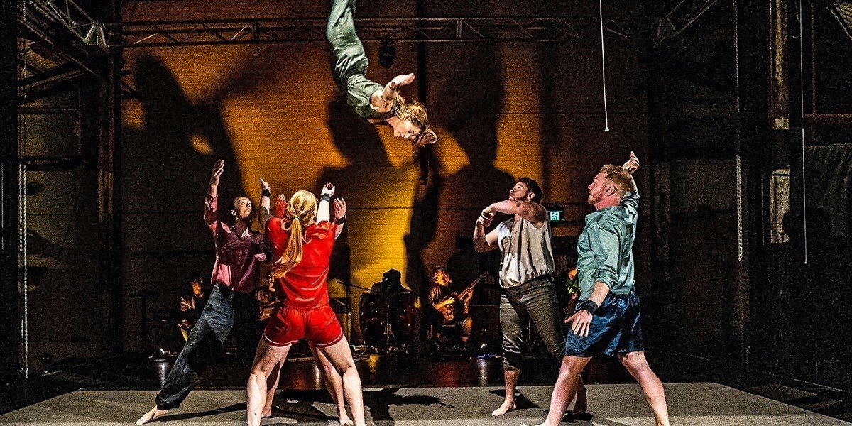 Six people on a stage performing. One person has been thrown into the air whilst the other five people are ready to catch them.  They are wearing an assortment of colours including red, maroon, grey, green & blue. There is yellow/orange/brown lighting in the background.