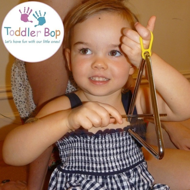 Toddler Bop - A photo of a young girl holding a triangle musical instrument. She is wearing a black and white gingham patterned top. The logo in the top corner features white circle with a purple and blue hand print with text that reads ‘Toddler Bop’ and ‘Let’s have fun with our little ones!’ in purple, blue and orange font.