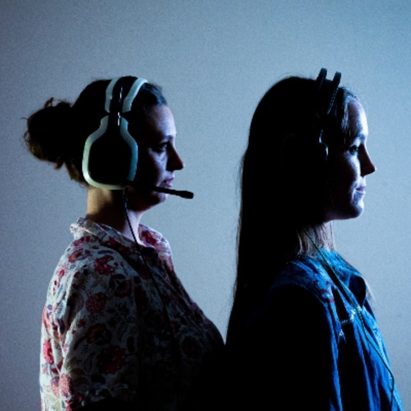 Two young women stand in darkness next to each other. They have a blank look on their face and headphones around their ears.