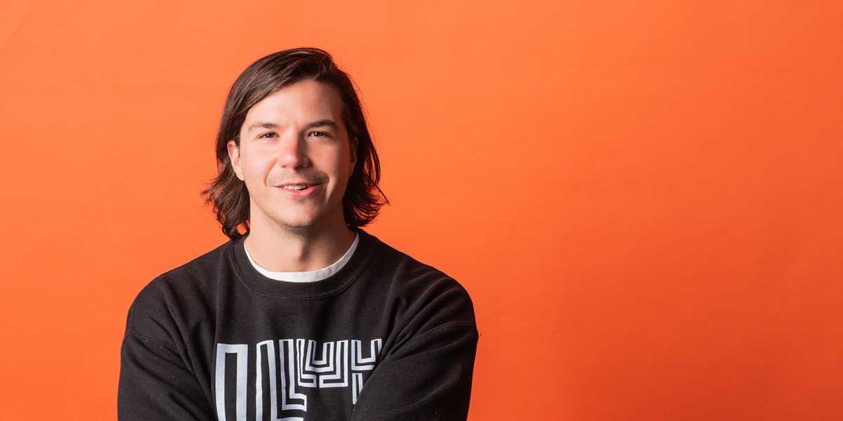 Luka wearing a black crewneck jumper and smiling in front of an orange background.