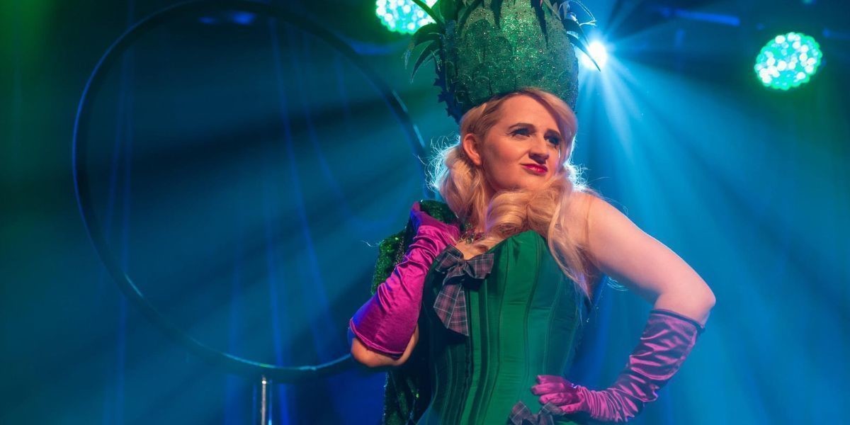 Peachy Pear stands on stage with blue lights behind her. She wears a greet corset with purple tartan bows, purple gloves and a head pieces made to look like a thistle. She is blonde. she her right hand over her right shoulder.