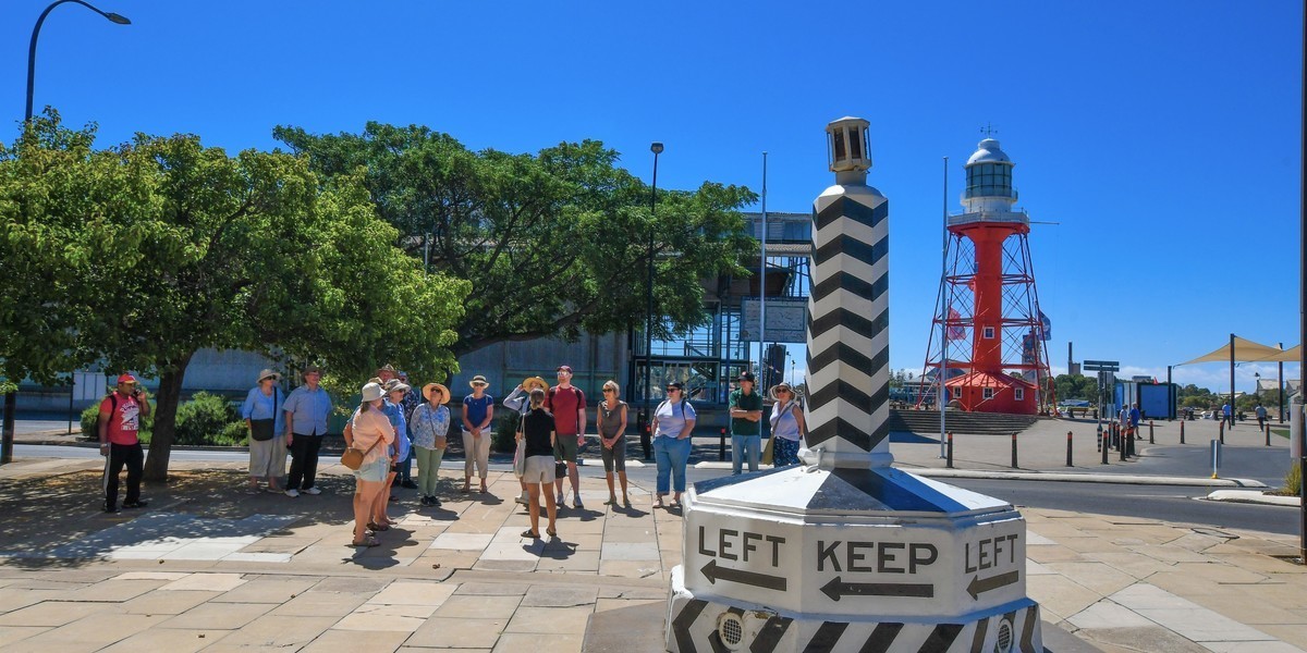 Port Adelaide Walking Tour - A tour guide speaks to a visitor group, with a lighthouse in the background and monument in the foreground.