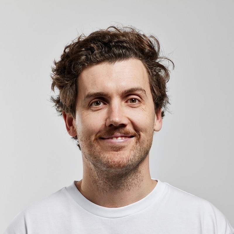 Daniel Connell sits looking at the camera, he's wearing a white shirt and is smiling. It is a portrait image taken from the shoulders up.