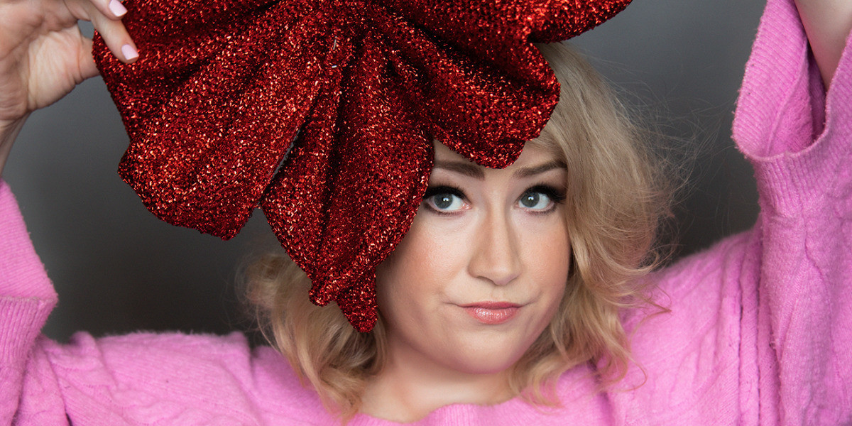 Rosie Waterland wears a pink top and a massive red sequinned bow on her head. Her arms are above her head holding the bow on each side and she wears a quizzical look as her eyes look up at the bow.