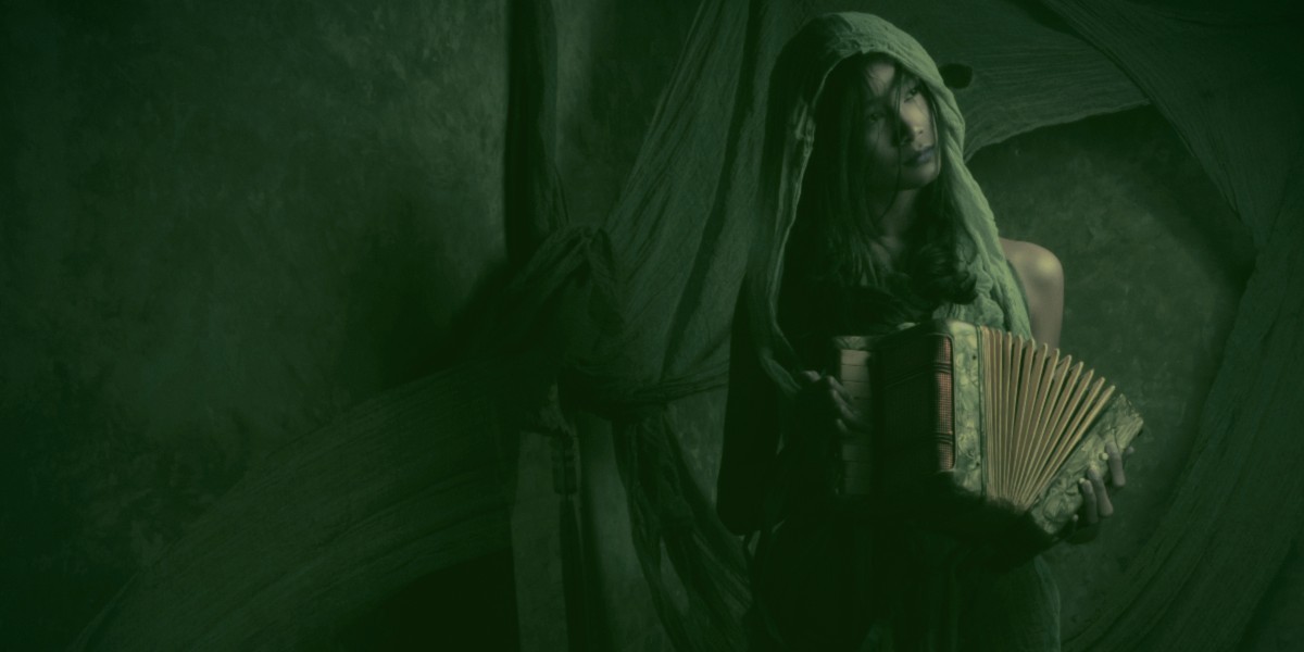 A girl wearing a hooded garment plays the accordion in the dim light.