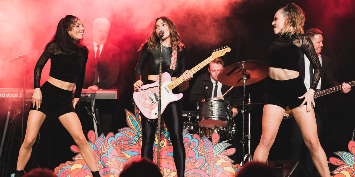 Three females in black costuming, standing on stage in the middle of a song and routine. Centre performing is playing a pink electric guitar behind a golden microphone stand