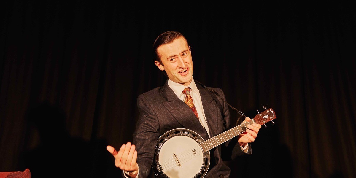 A young man dressed in an old-school dark zoot suit with a white shirt and bright, paisley tie holds a banjo ukulele with one hand while the other gestures to the audience with a big grin.