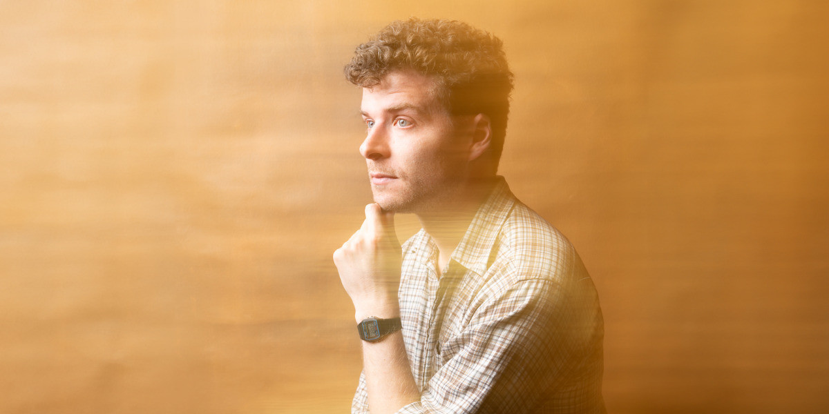 Oliver is standing on an angle giving a side profile. His hand is resting underneath his chin. He has short curly hair and blue eyes. He is wearing a brown checked shirt. The image has a blurry texture across the image.