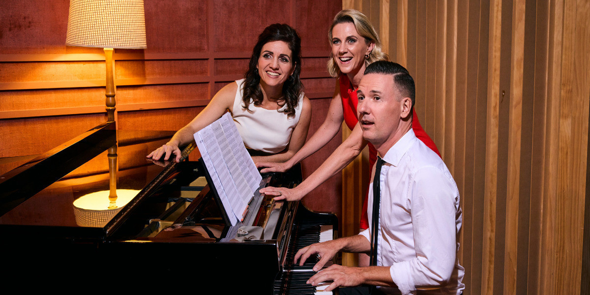 A man with short brown hair sits and plays piano, wearing a white shirt and black slim tie. A blonde woman stands behind him wearing an orange dress, leaning on the front of the piano, while a shorter brunette woman wearing a white top leans on the back of the piano. There is sheet music on the music stand, a lamp on in the background, and they all smile, looking slightly off camera as though someone has just entered the room they are rehearsing in.