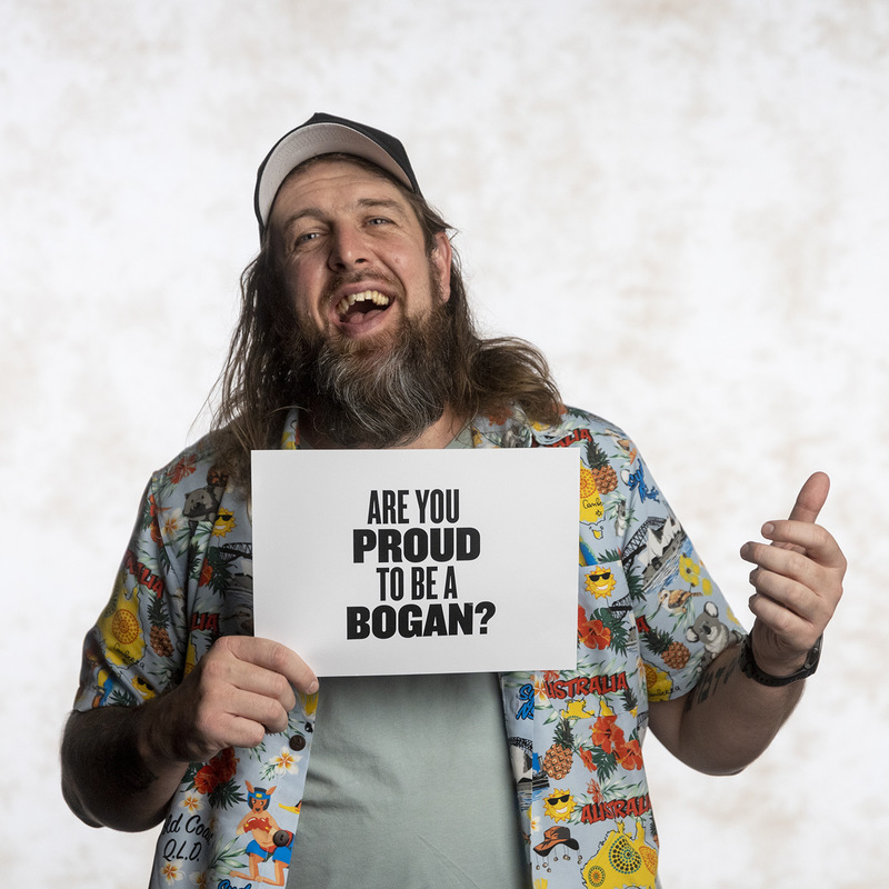 A hairy man, with a long mullet, beard wearing a hat and a colourful shirt. He is holding a piece of paper saying “ are you proud to be a Bogan?”