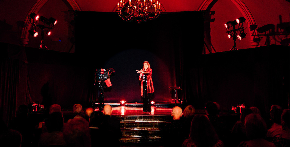 Stephanie Rummel standing on stage, lit by red light with a crowd before her.