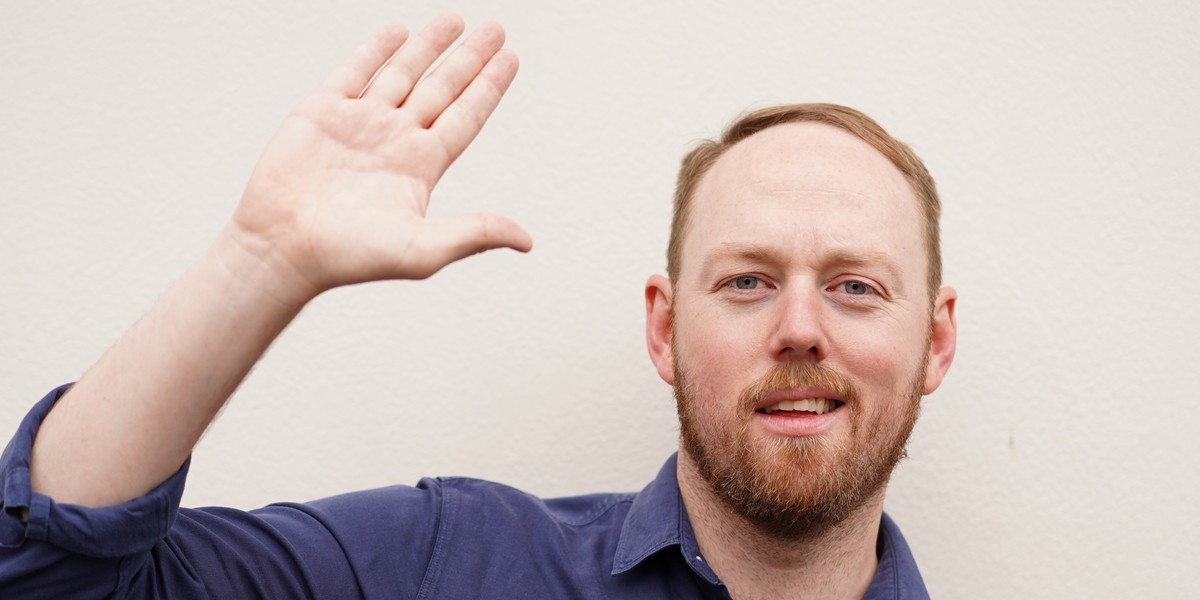 A photograph of a man with short strawberry blonde hair and beard smirking at the camera with one hand in the air, as if to wave. The background is a cream colour.
