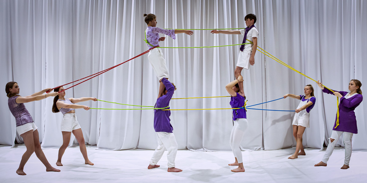Four people pulling on ropes attached to two people standing on the shoulders for two other people.